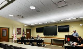 training room showing 3 monitors, 1 man & 2 women at a table, a man at the podium and another woman sitting by herself at a tabl